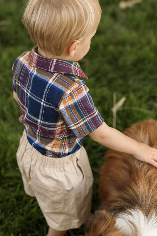 A young child wearing the Ivy City Co Baby Boy James Romper in Midnight Plaid stands on grass, gently petting a brown and white dog. The photo is taken from above and behind them.