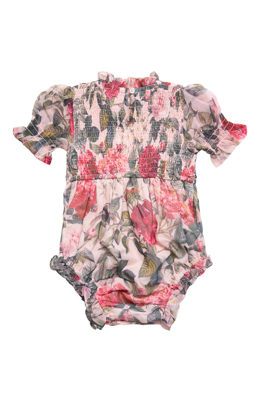 The Ivy City Co Baby Cordelia Romper in Pink Bloom features a floral print, short puff sleeves, smocked bodice, ruffled leg openings, and diaper snaps in soft pink, green, and white.