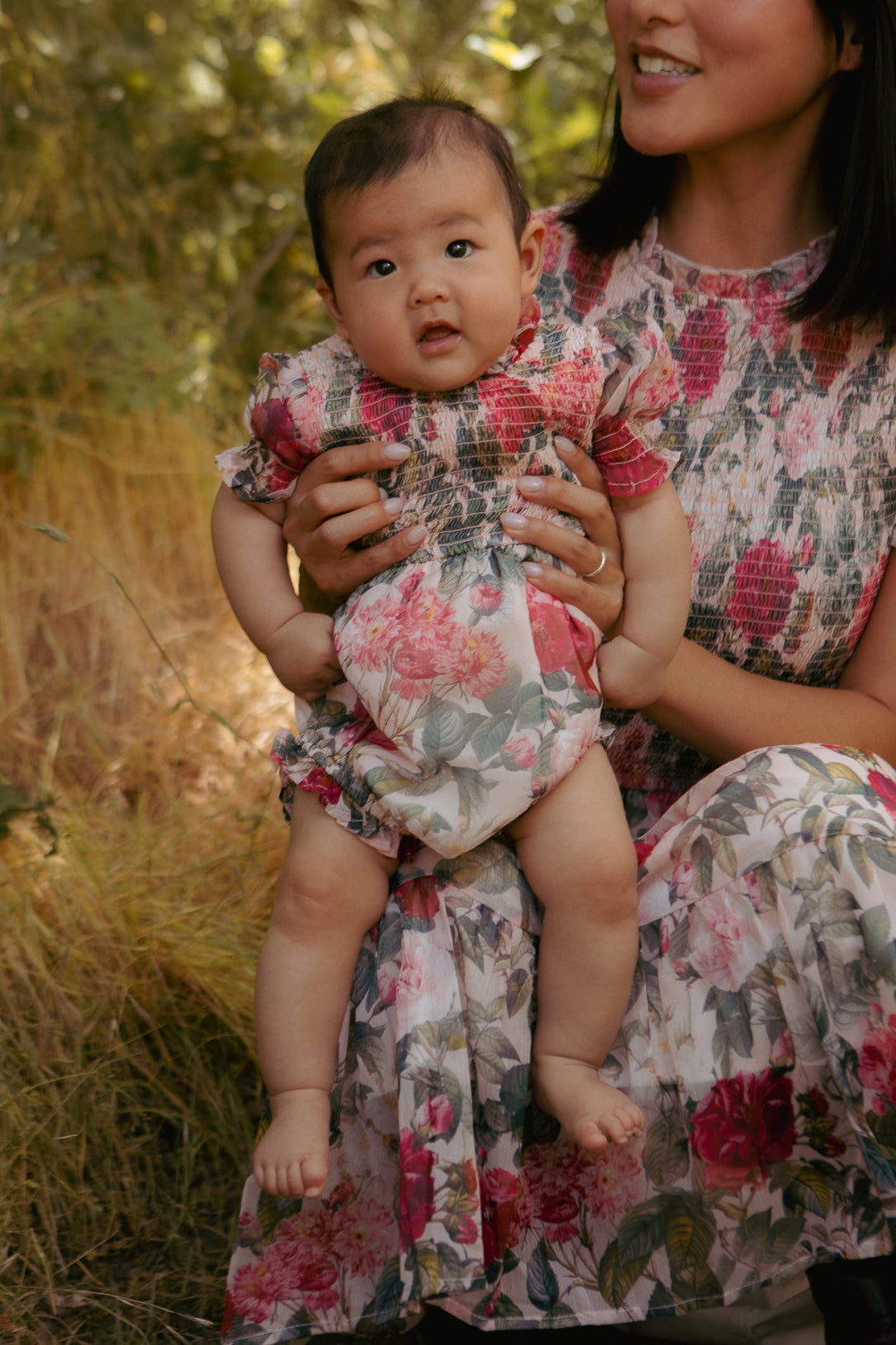 A woman in a floral dress holds a baby outdoors among tall grass. The baby, wearing the Ivy City Co Baby Cordelia Romper in Pink Bloom with diaper closure snaps, looks at the camera while the woman smiles.
