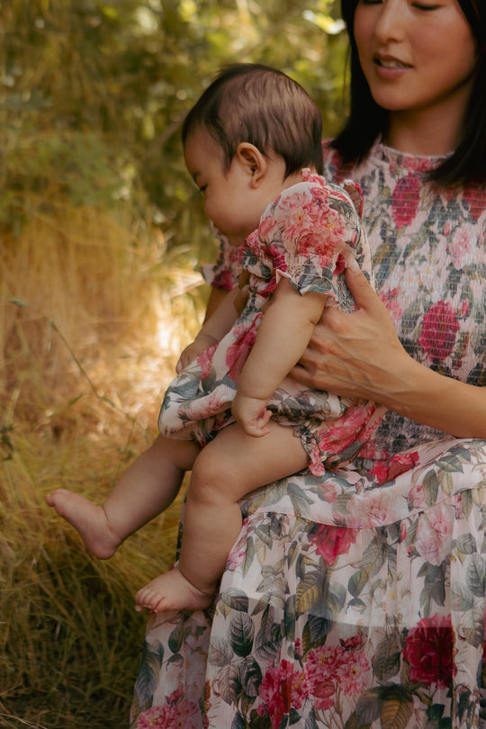A woman in a floral dress holds a baby wearing the Ivy City Co Baby Cordelia Romper in Pink Bloom, featuring convenient diaper closure snaps, as they sit outdoors amid greenery and tall golden grass.