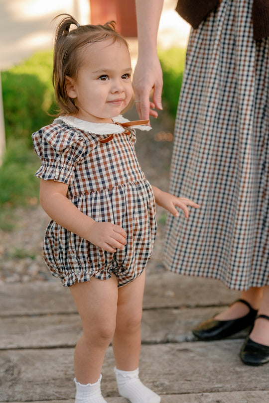 A toddler wearing the Ivy City Co Baby Emery Romper in Chestnut Gingham stands on wooden boards, holding hands with an adult who is mostly out of frame and dressed in a matching plaid skirt. Both are outdoors, and the child smiles softly.
