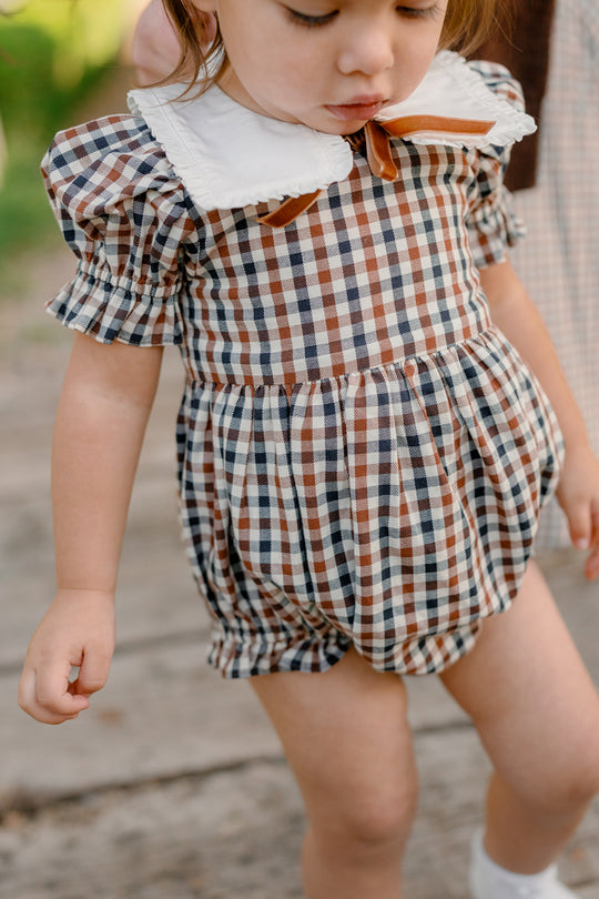 A toddler stands outdoors on a wooden path wearing the Ivy City Co Baby Emery Romper in Chestnut Gingham, featuring short puffed sleeves, a crisp white collar, and a bow in charming brown, black, and white checkered cotton.