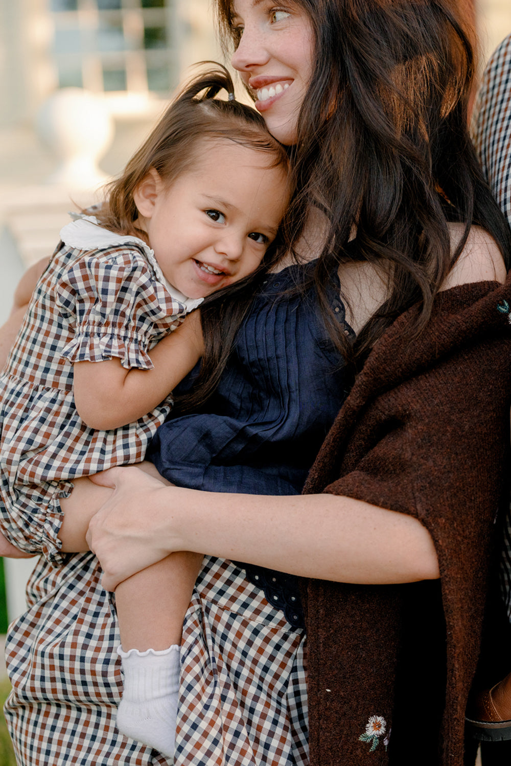 A smiling woman in a dark dress holds a young girl dressed in the Ivy City Co Baby Emery Romper in Chestnut Gingham. The child hugs her, beaming with happiness, while the woman gazes at her lovingly. They are outdoors, bathed in gentle sunlight.