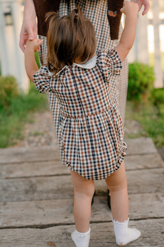 A toddler dressed in the Ivy City Co Baby Emery Romper in Chestnut Gingham holds hands with an adult—also wearing plaid—while standing on a wooden path outside, surrounded by green grass and a white fence under bright daylight.