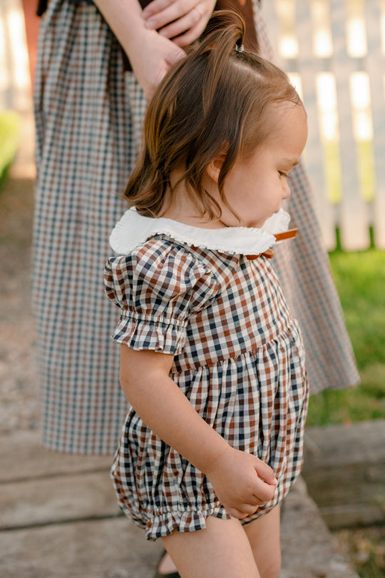 A young child wears the Ivy City Co Baby Emery Romper in Chestnut Gingham while standing outdoors and holding an adult's hand. The adult, partially visible and also dressed in plaid, stands beside the child against a backdrop of green grass and a white fence.