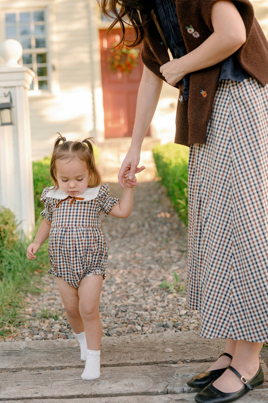 A toddler wearing the Ivy City Co Baby Emery Romper in Chestnut Gingham with a matching bow holds hands with a woman in a plaid skirt as they walk outside on a stone path near a house with a red door.