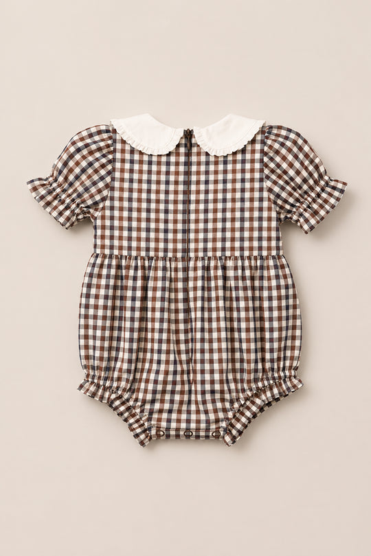 The Baby Emery Romper in Chestnut Gingham by Ivy City Co is a brown and white gingham cotton romper featuring short puff sleeves with ruffled edges, a white ruffled collar, a back zipper, and a snap-bottom closure. This adorable romper is shown flat against a solid light beige background.