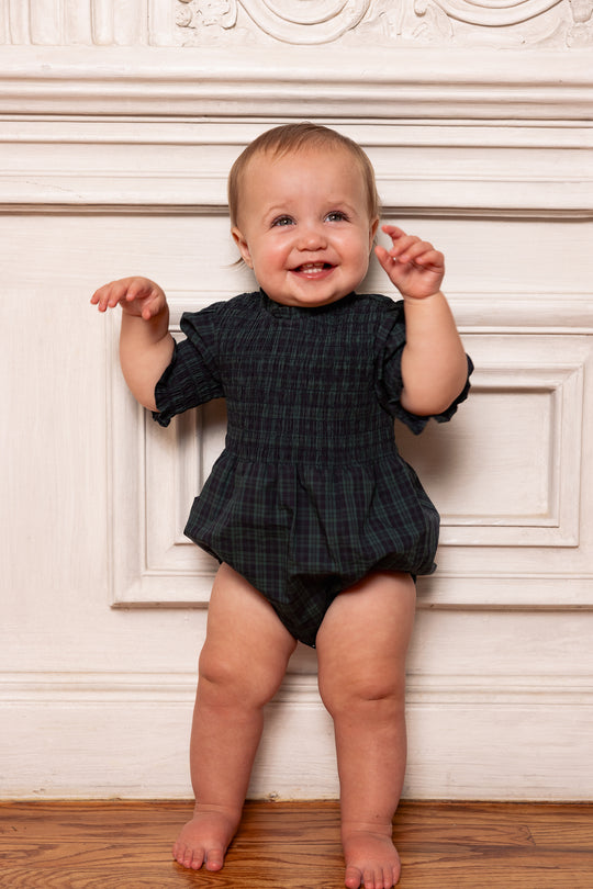 Baby in a dark checkered romper standing against a decorative wall.