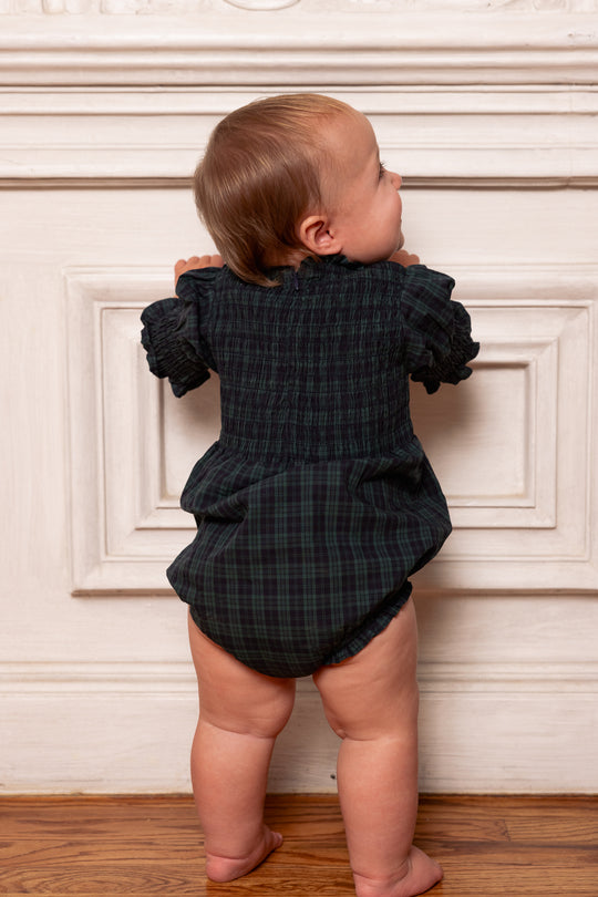 Baby in a dark plaid romper standing against a white wall.