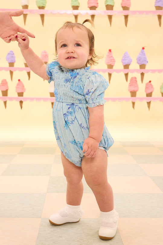 Baby in a blue floral romper standing on a checkered floor with ice cream cone decorations in the background.