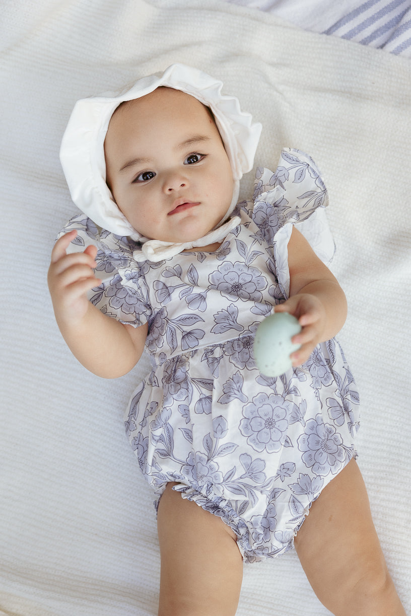 A baby wearing the Ivy City Co Baby Hattie Romper in Blue Floral lies on a soft white blanket, looking up and holding a small, pale blue egg-shaped object.