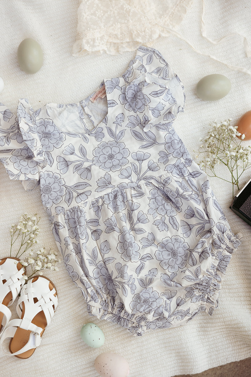 The Ivy City Co Baby Hattie Romper in Blue Floral is displayed on a textured white blanket, surrounded by pastel Easter eggs, white sandals, delicate white flowers, and a piece of lace fabric.