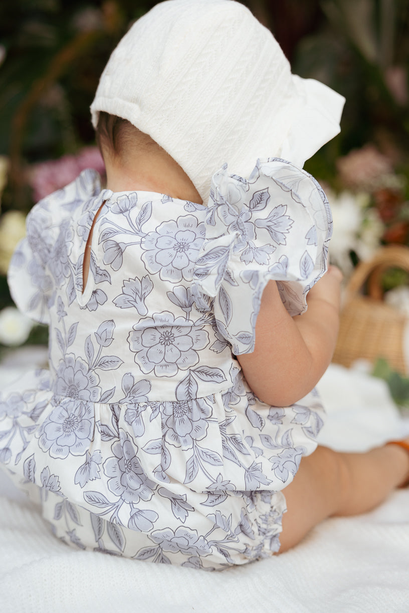 A baby wearing the Ivy City Co Baby Hattie Romper in Blue Floral and a white bonnet sits on a white blanket, facing away, with softly blurred green plants and wicker decor in the background.