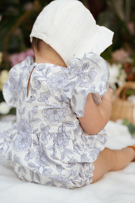 A baby wearing the Ivy City Co Baby Hattie Romper in Blue Floral and a white bonnet sits on a white blanket, facing away, with softly blurred green plants and wicker decor in the background.
