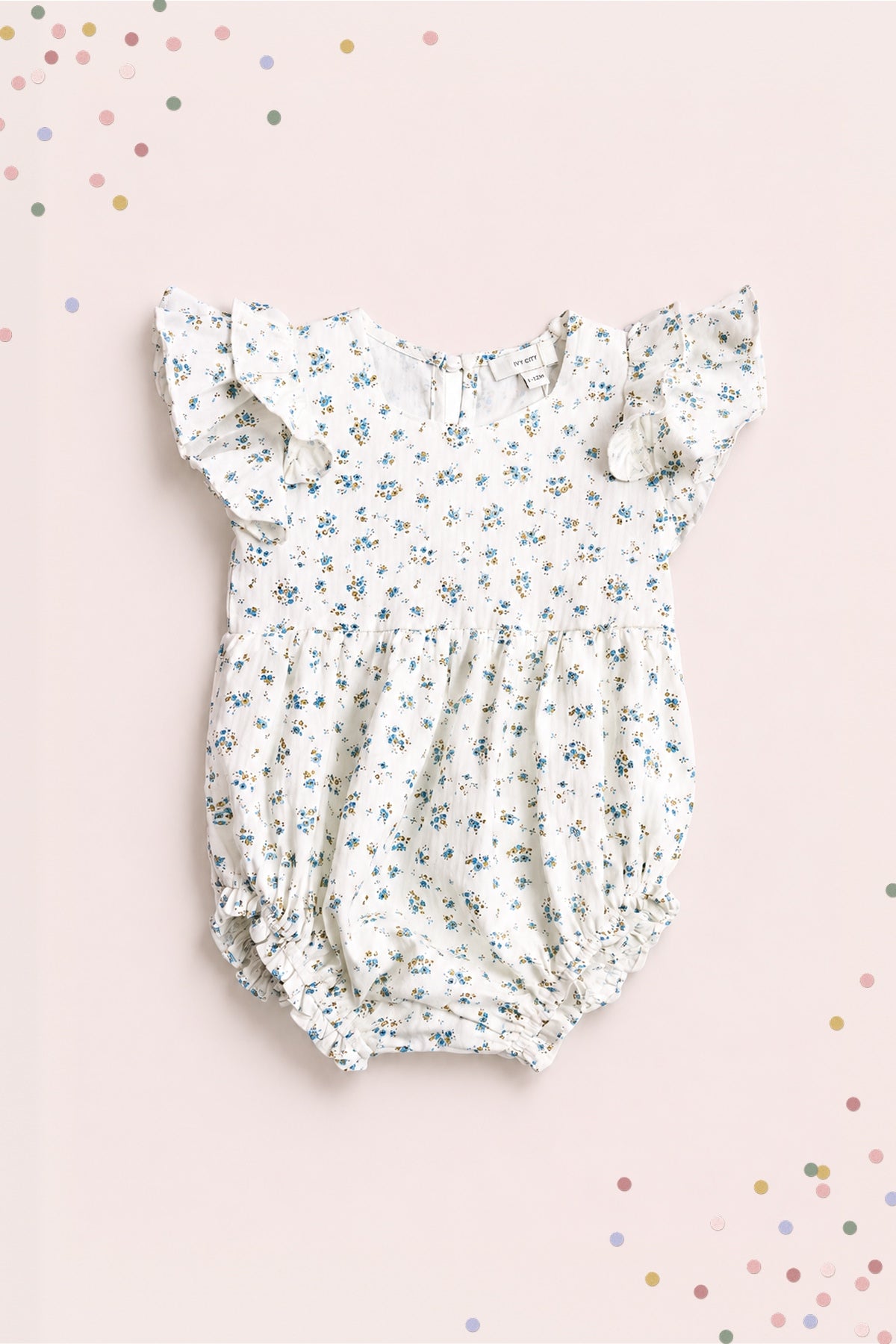 The Ivy City Co Baby Hattie Romper in Madeline Blue features a white cotton fabric with blue floral print, ruffled short sleeves, and elastic leg openings—perfect for special occasions. Shown on a light pink background with colorful confetti dots.