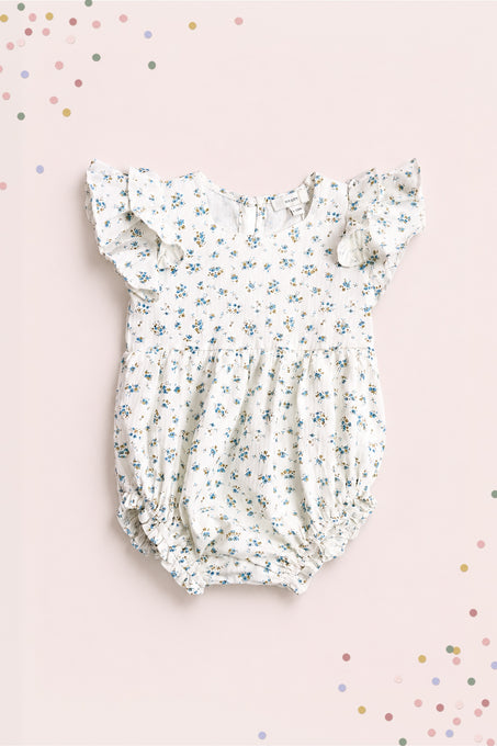 The Ivy City Co Baby Hattie Romper in Madeline Blue features a white cotton fabric with blue floral print, ruffled short sleeves, and elastic leg openings—perfect for special occasions. Shown on a light pink background with colorful confetti dots.