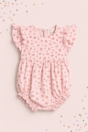 The Ivy City Co Baby Hattie Romper in Madeline Pink features ruffled sleeves and a floral print, displayed on a matching background with pastel confetti—an adorable first birthday outfit.