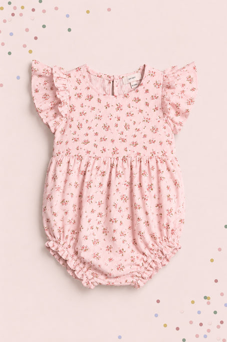 The Ivy City Co Baby Hattie Romper in Madeline Pink features ruffled sleeves and a floral print, displayed on a matching background with pastel confetti—an adorable first birthday outfit.