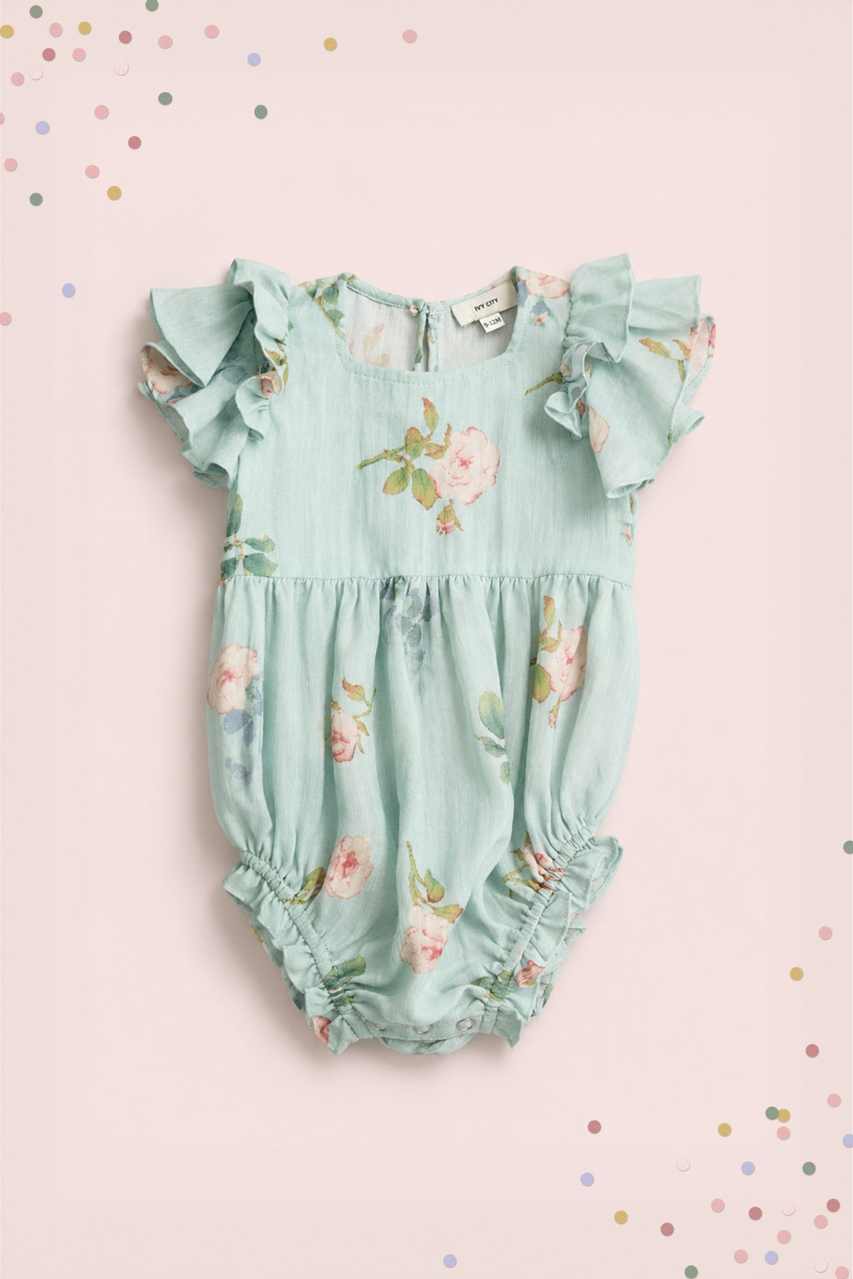 The Ivy City Co Baby Hattie Romper in Roselyn Blue features a light blue cotton-linen fabric with pink floral print, flutter sleeves, and gathered leg openings, pictured on a pale pink background with colorful confetti dots.