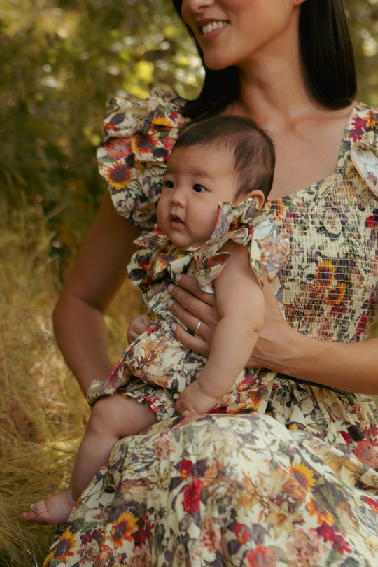 A woman in a floral dress holds a smiling baby wearing the Ivy City Co Baby Hattie Romper in Rose Vine, featuring easy diaper snaps, as they relax together outdoors surrounded by lush greenery.
