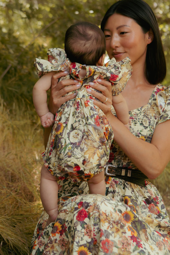 A woman sits outdoors holding a baby, both dressed in Ivy City Cos Rose Vine print. The baby wears the Baby Hattie Romper in Rose Vine, featuring convenient diaper snaps. Lush greenery and tall grass complete this warm, natural scene.