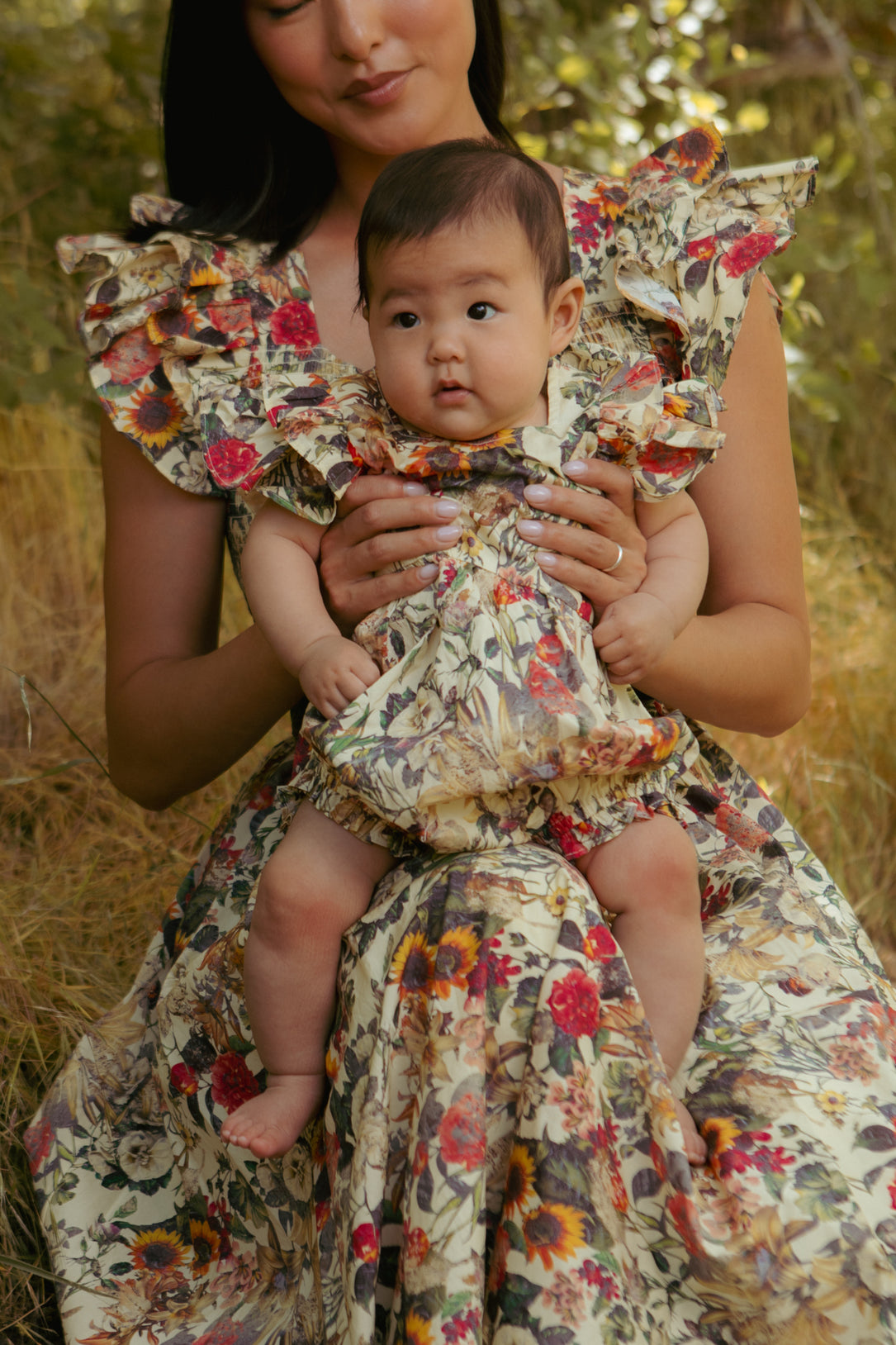 A woman sits outdoors with her baby on her lap, both in Ivy City Co’s Rose Vine print. The baby wears the Baby Hattie Romper in Rose Vine, featuring diaper snaps and ruffled sleeves, surrounded by greenery and tall grass.