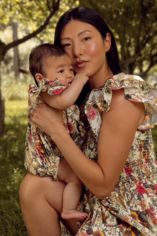 A woman holds a baby outdoors, both in matching Ivy City Co Rose Vine outfits. The baby wears the Hattie Romper, featuring convenient diaper snaps. They stand close together amid green trees and gentle sunlight.