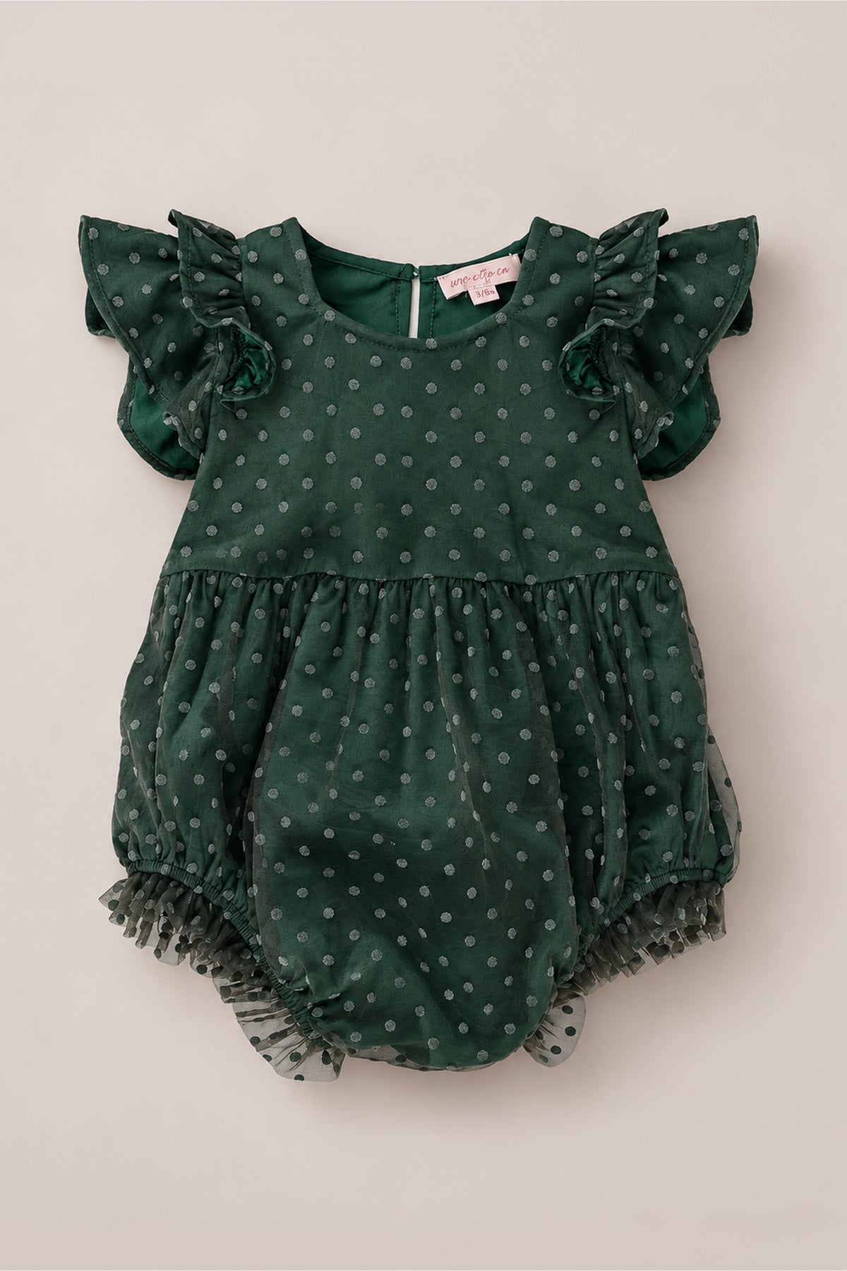 The Ivy City Co Baby Hattie Romper in Green Flocked Dot Tulle features a dark green hue with short ruffled sleeves, a sheer polka-dotted green flocked tulle overlay, and ruffled trim at the leg openings. A tag is sewn into the inside of the collar. Set against a light beige background, this romper is perfect for family photos.