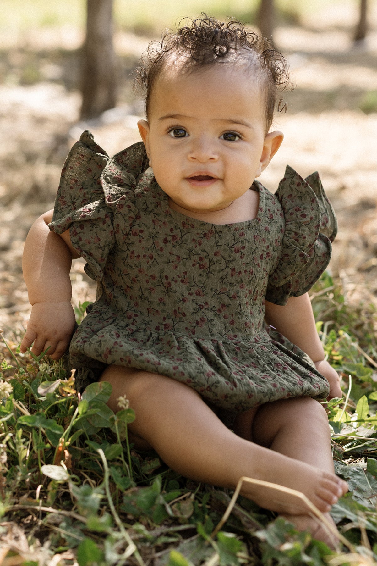 A baby with curly hair sits on the grass outdoors, looking at the camera with a gentle smile, dressed in the Ivy City Co Baby Hattie Romper in Woodland Floral featuring charming flutter sleeves. Sunlight filters through the trees in the background.