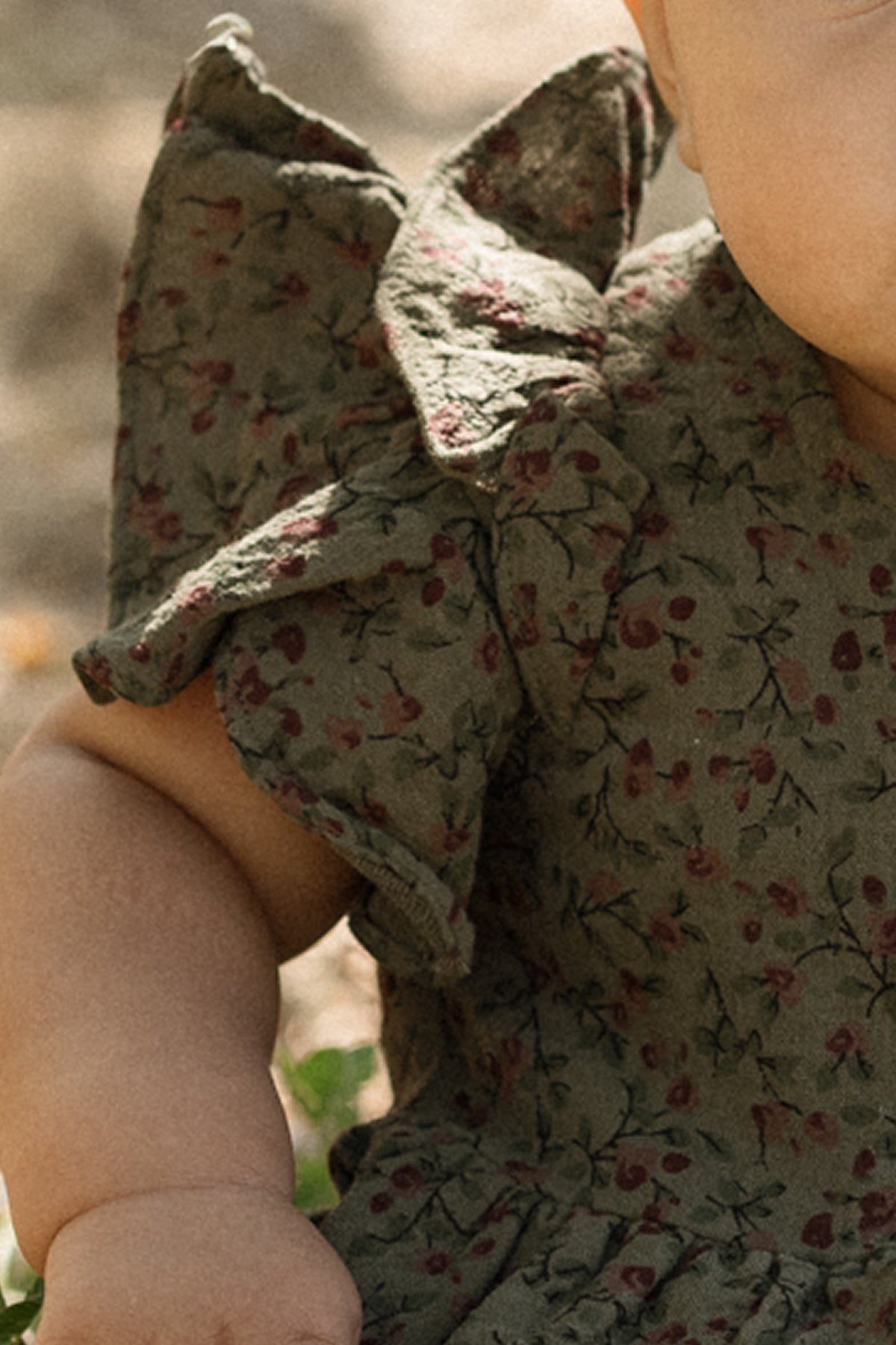 A close-up of a baby wearing the Ivy City Co Baby Hattie Romper in Woodland Floral, a green cotton romper adorned with a petite floral print and charming flutter sleeves, with the baby's arm and part of their face visible.