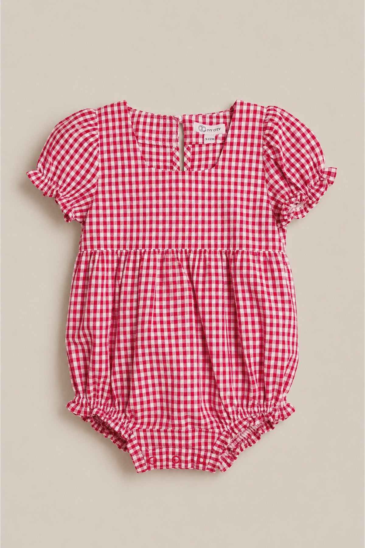 The Baby Jessie Romper in Red Gingham by Ivy City Co features short puff sleeves, ruffled edges, a gathered waist, back buttons, and elastic leg openings—ideal for the Fourth of July. Shown laid flat on a neutral background.