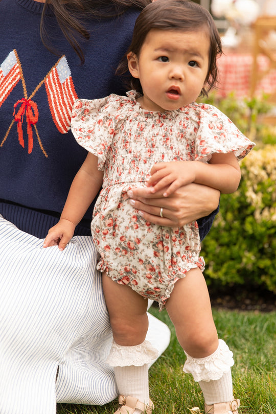 A toddler wears the Ivy City Co Baby Jovie Romper in Ivory Heritage Rose and ruffled socks, held by an adult in a navy sweater with American flags and a red bow. They're outside on grass, greenery blurred behind—a perfect Mom and Me matching moment.