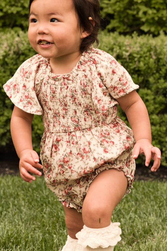 A young child in the Ivy City Co Baby Jovie Romper in Ivory Heritage Rose and cream socks plays on green grass with bushes behind, smiling—perfect for adorable Mom and Me matching moments.
