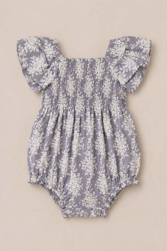The Ivy City Co Baby Lisa Romper in Jolie Fleur features flutter sleeves, a light purple hue adorned with a white floral pattern, and gathered fabric accents at the shoulders, chest, and leg openings. A charming smocked bodice adds texture and elegance to this adorable piece.