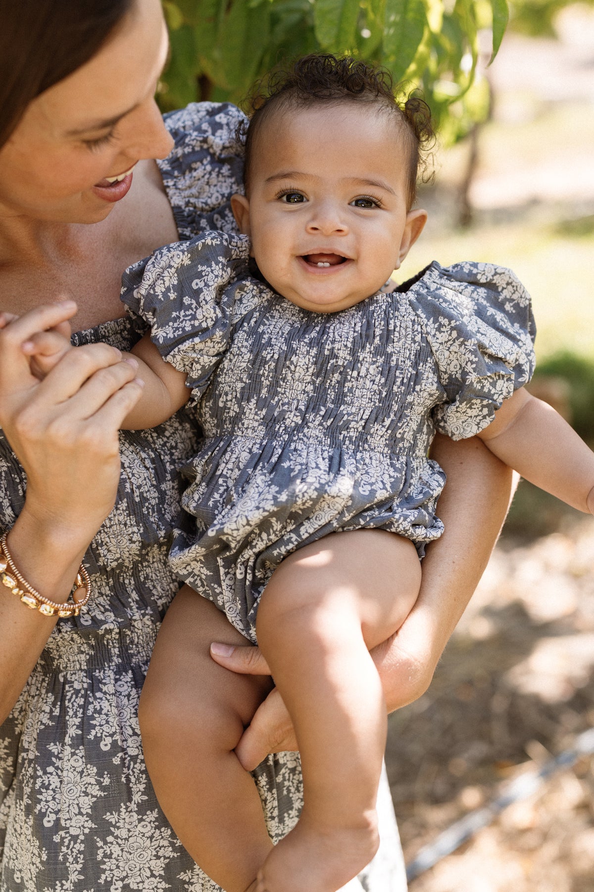 A smiling baby wears the Ivy City Co Baby Lisa Romper in Jolie Fleur, featuring a smocked bodice and puff sleeves, while being held by an adult in a matching dress. They are outdoors with greenery in the background.