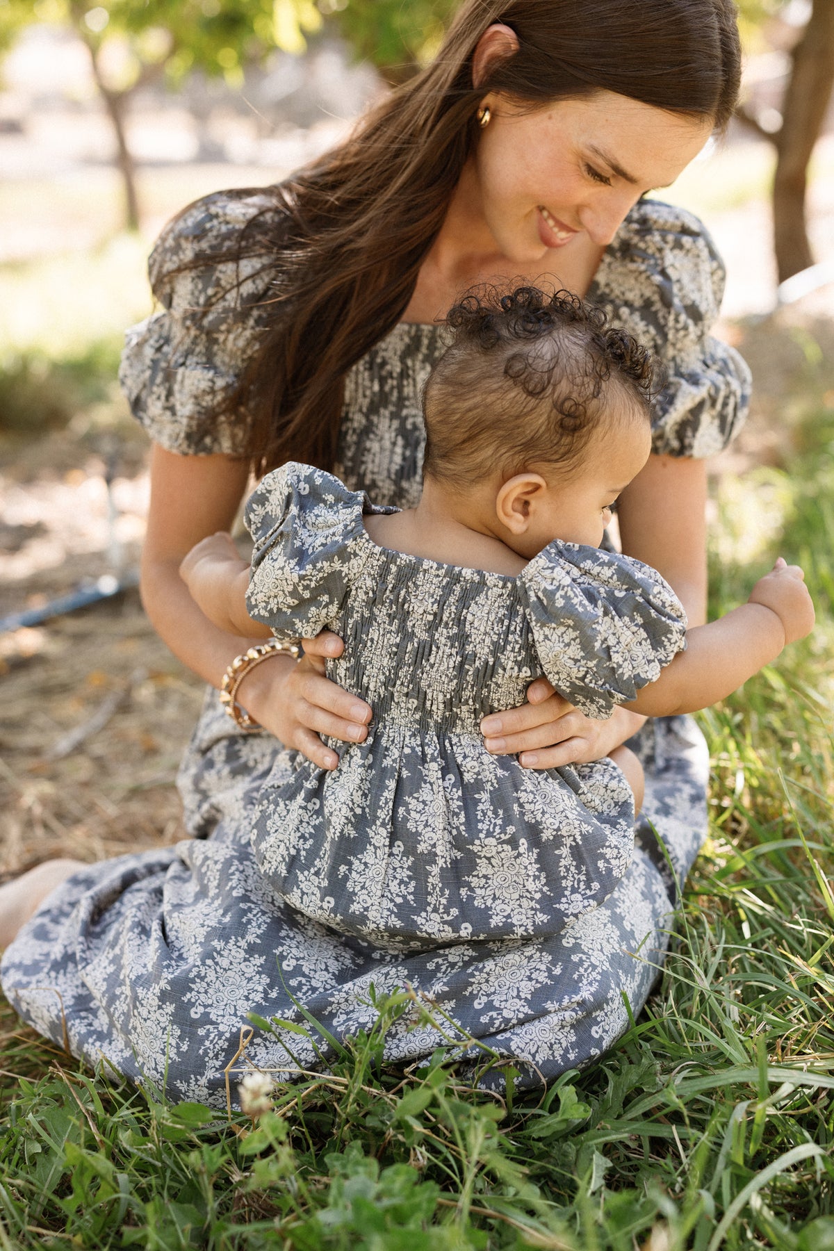 A woman and her baby wearing matching floral outfits sit on the grass outdoors. The woman’s dress features a delicate smocked bodice and puff sleeves, while the baby is dressed in the adorable Ivy City Co Baby Lisa Romper in Jolie Fleur. Smiling, the woman gently hugs her baby, who looks down and reaches toward the ground as sunlight filters through the trees around them.