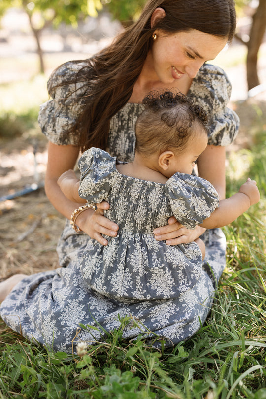 A woman and her baby wearing matching floral outfits sit on the grass outdoors. The woman’s dress features a delicate smocked bodice and puff sleeves, while the baby is dressed in the adorable Ivy City Co Baby Lisa Romper in Jolie Fleur. Smiling, the woman gently hugs her baby, who looks down and reaches toward the ground as sunlight filters through the trees around them.