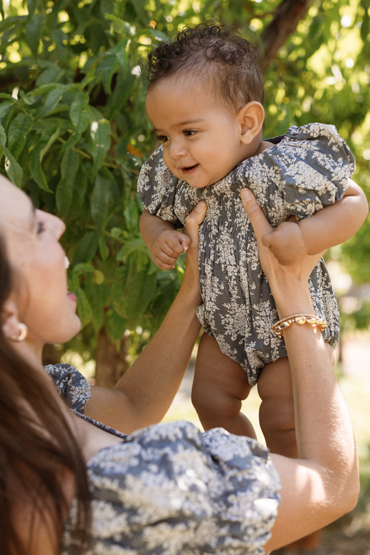 A woman lifts a smiling baby in her arms outdoors, both dressed in matching blue floral outfits with puff sleeves as green leaves from a tree create a lush backdrop. The baby’s adorable look is completed by the Ivy City Co Baby Lisa Romper in Jolie Fleur, featuring a delicate smocked bodice.