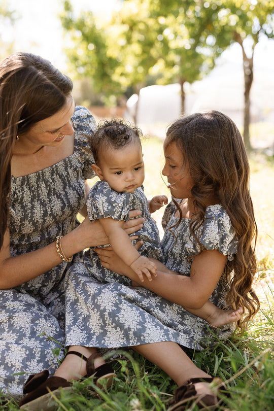A woman and two young children sit on grass outdoors, all wearing matching gray and white floral dresses with puff sleeves and smocked bodices. The woman holds the baby, who is dressed in the adorable Ivy City Co Baby Lisa Romper in Jolie Fleur, while the older child smiles at the baby. Sunlight filters through green trees in the background.