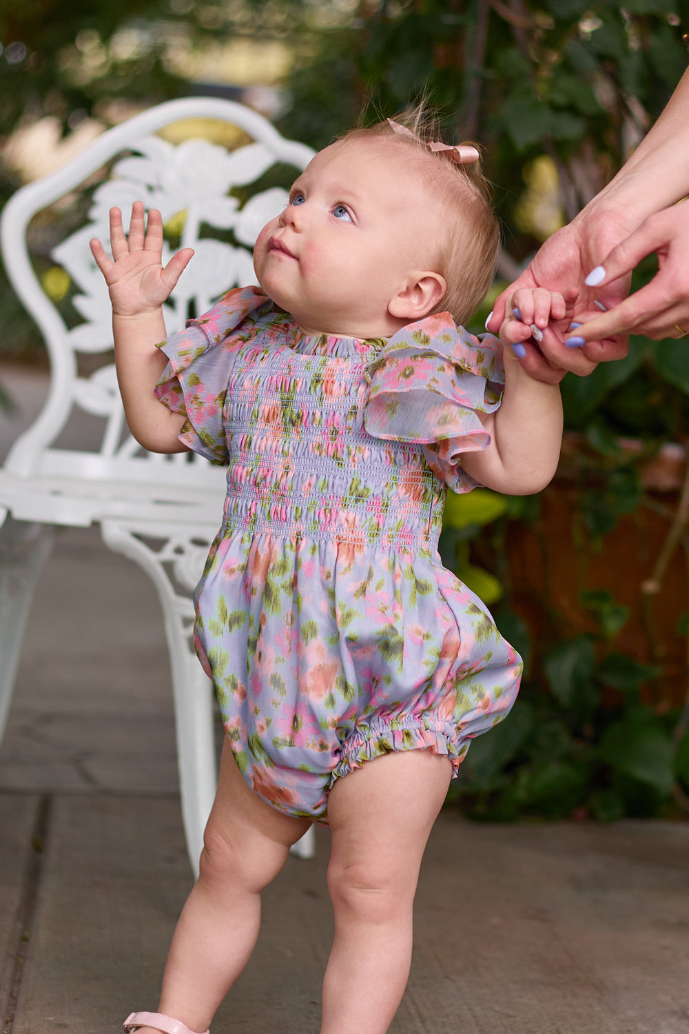 A toddler wears the Ivy City Co Baby Lottie Romper in Vibrant English Garden while holding an adult’s hand on a patio, near a white chair and lush greenery—a charming garden-inspired baby outfit moment.