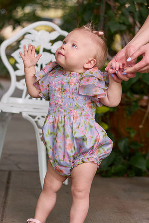 A toddler wears the Ivy City Co Baby Lottie Romper in Vibrant English Garden while holding an adult’s hand on a patio, near a white chair and lush greenery—a charming garden-inspired baby outfit moment.
