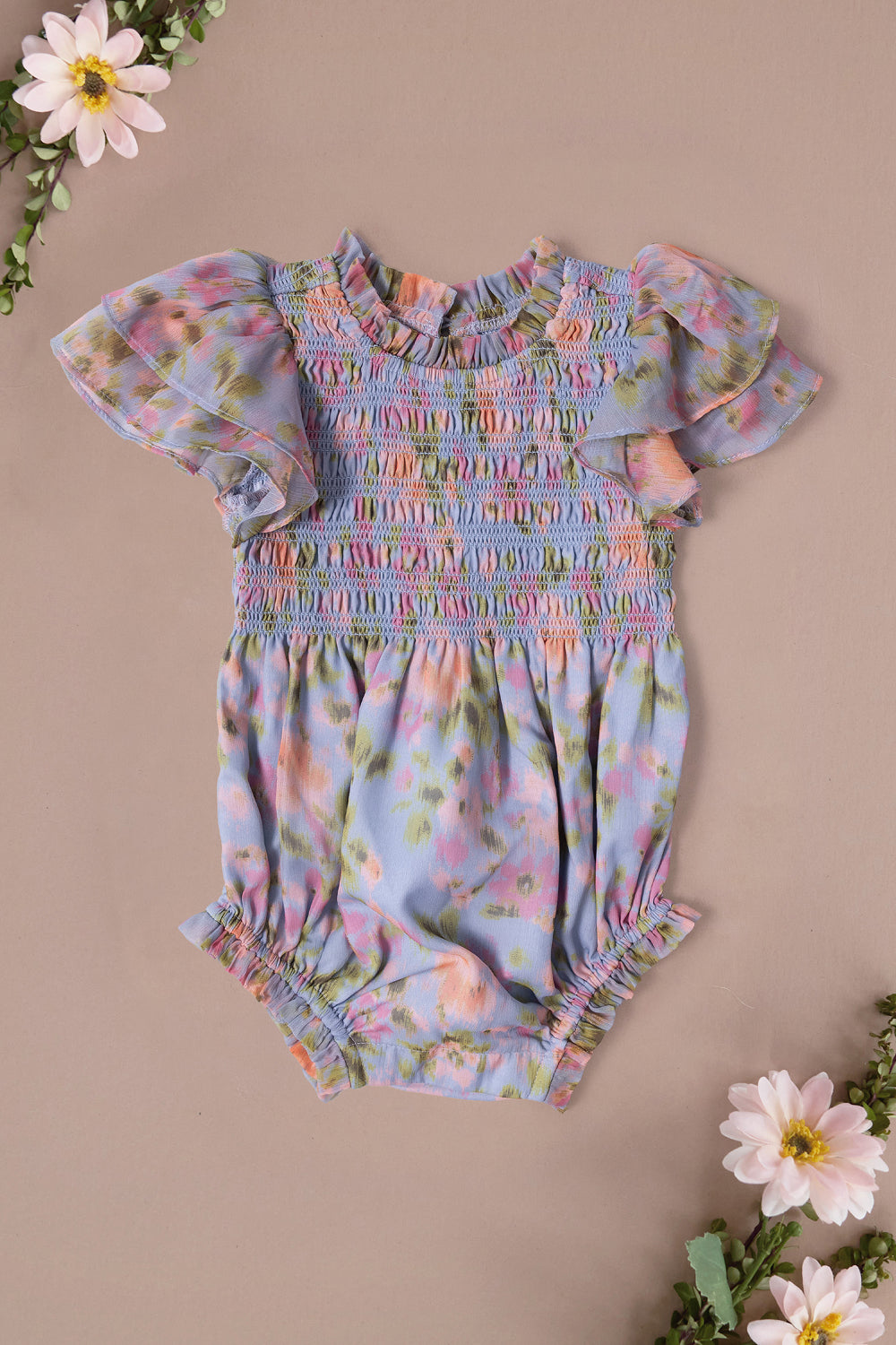 The Ivy City Co Baby Lottie Romper in Vibrant English Garden features pastel florals, short ruffled sleeves, and gathered hems. Displayed on a light beige background with pink and white daisies—perfect for a charming garden-themed look.