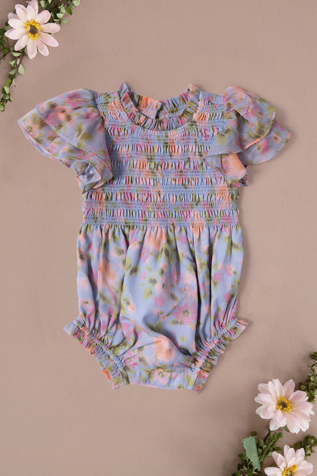 The Ivy City Co Baby Lottie Romper in Vibrant English Garden features pastel florals, short ruffled sleeves, and gathered hems. Displayed on a light beige background with pink and white daisies—perfect for a charming garden-themed look.