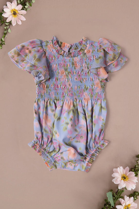The Ivy City Co Baby Lottie Romper in Vibrant English Garden features pastel florals, short ruffled sleeves, and gathered hems. Displayed on a light beige background with pink and white daisies—perfect for a charming garden-themed look.