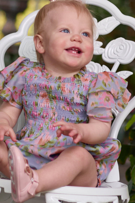 A smiling baby with blonde hair sits on a white ornate chair, dressed in the Ivy City Co Baby Lottie Romper in Vibrant English Garden and pink shoes, surrounded by greenery.