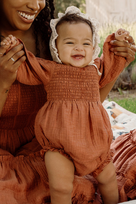 A smiling baby in the Ivy City Co Baby Madeline Romper in Apple Cider, featuring puff sleeves and a bonnet, is held up by an adult wearing a matching dress while outdoors on a blanket.