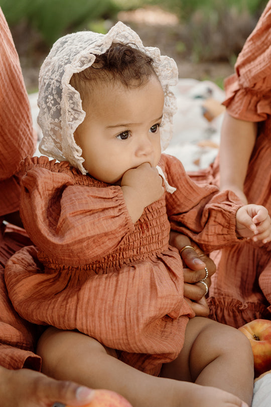 A baby dressed in the Ivy City Co Baby Madeline Romper in Apple Cider, featuring a smocked bodice and puff sleeves and paired with a lace bonnet, sits outdoors sucking on their fingers while being gently held by an adult.