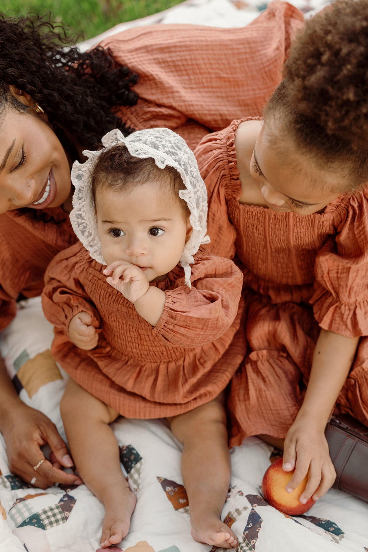 A baby wearing the Ivy City Co Baby Madeline Romper in Apple Cider, featuring puff sleeves and a smocked bodice, sits on a quilt while sucking their thumb. Beside the baby are a woman and an older child dressed in matching outfits; the child holds an apple.