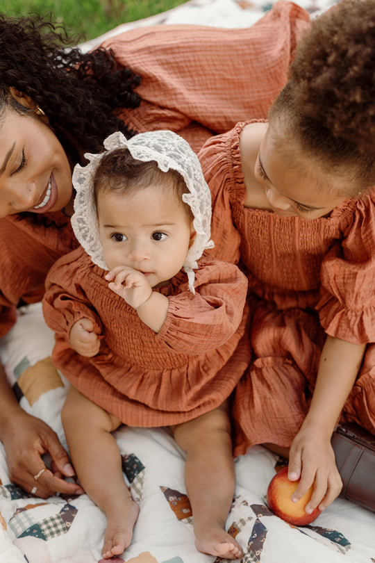A baby wearing the Ivy City Co Baby Madeline Romper in Apple Cider, featuring puff sleeves and a smocked bodice, sits on a quilt while sucking their thumb. Beside the baby are a woman and an older child dressed in matching outfits; the child holds an apple.