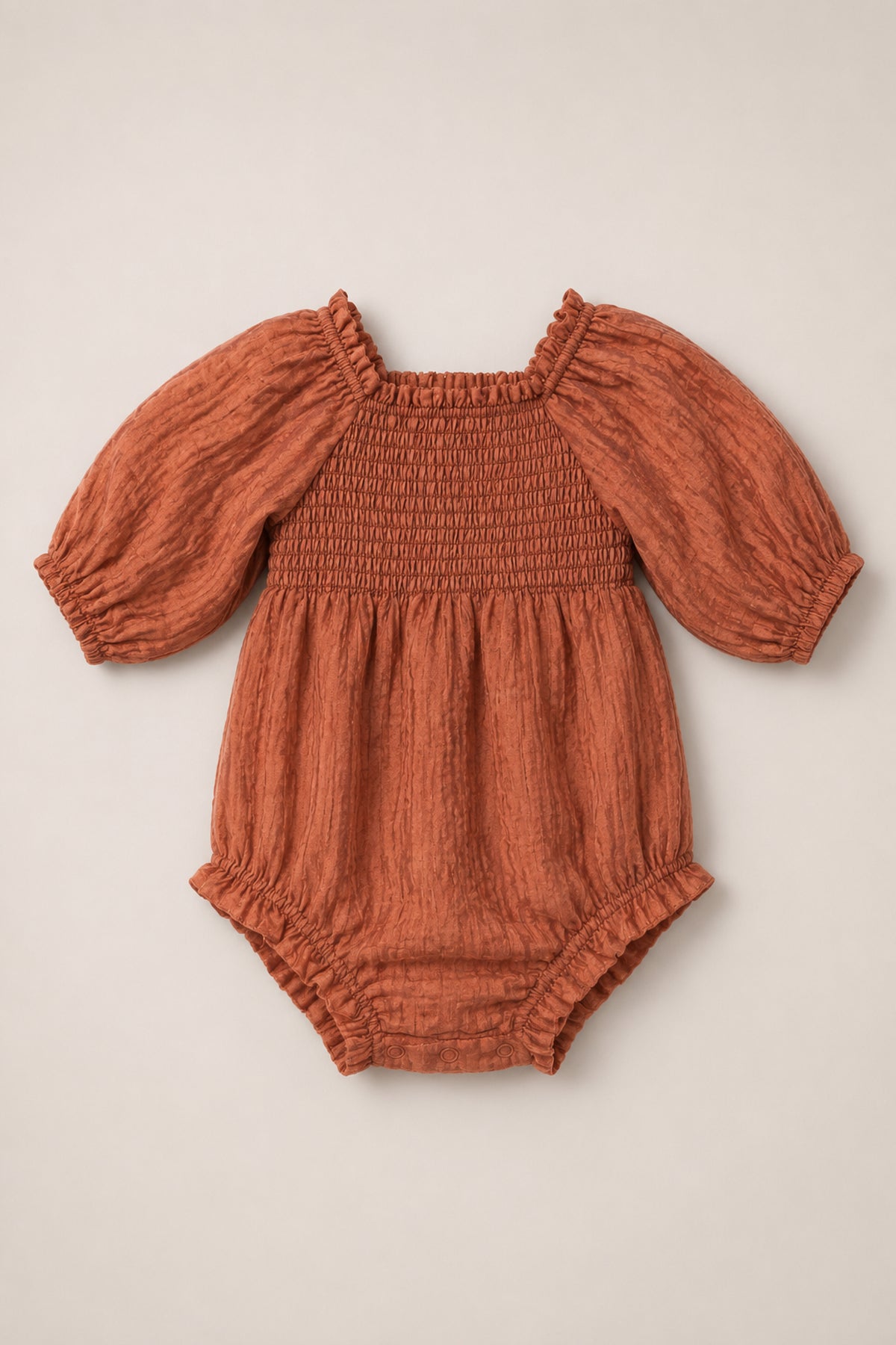 Ivy City Co’s Baby Madeline Romper in Apple Cider features a rust-colored long-sleeve design with a smocked bodice, subtle puff sleeves, elastic cuffs, and gathered leg openings, displayed on a neutral background.